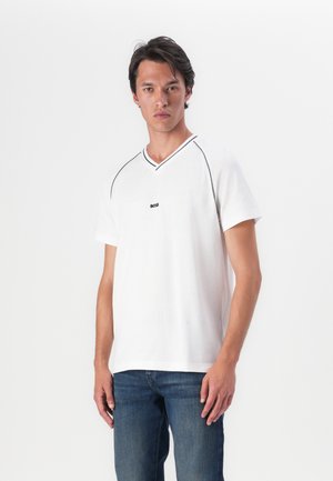 T-shirt blanc à manches courtes avec un col en V, tissu texturé, accents de couture noirs et un petit logo sur la poitrine. Porté avec un jean bleu.