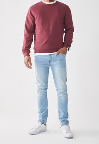 Sudadera granate con cuello redondo y detalle de logo negro, combinada con jeans ajustados azul claro y zapatillas blancas. Diseño sencillo y estilo casual.
