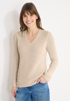Femme souriante aux cheveux bruns portant un pull en maille beige à col en V et un jean bleu, debout avec une main dans la poche devant un fond blanc.