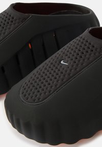 Μαύρα παπούτσια χωρίς κορδόνια με υφή στην επιφάνεια, διακοσμημένα με τρύπες στο πάνω μέρος και στρογγυλεμένη, διαμορφωμένη σόλα. Μικρή ασημί λεπτομέρεια λογότυπου ορατή.