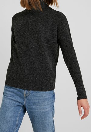 Person wearing a dark gray turtleneck sweater and blue jeans, shown from behind with one arm slightly extended.