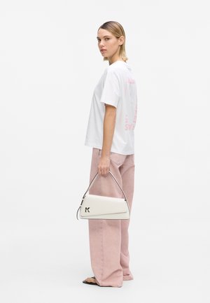 Femeie în tricou alb și pantaloni roz cu picioare largi, ținând o geantă albă cu logo-ul „K”, stând pe un fundal simplu.