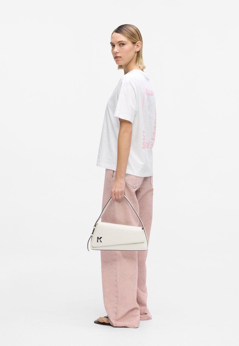 Γυναίκα με λευκό μπλουζάκι και ροζ παντελόνι φαρδιάς γραμμής, κρατώντας λευκή τσάντα με λογότυπο "K", στέκεται μπροστά σε ένα απλό φόντο.