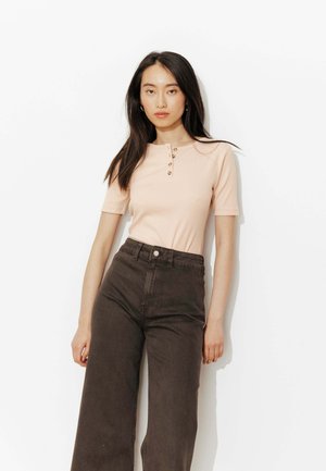 Jeune femme aux cheveux lisses et foncés portant un haut henley à manches courtes beige clair et un pantalon large taille haute couleur marron foncé.