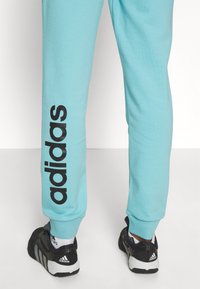 Ben klädda i ljusblå Adidas joggingsbyxor med stort svart logotyp på vänster ben och svarta Adidas sneakers med vita sulor.
