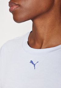 Vit T-shirt med rundad halsringning, med en blå Puma-logotyp på bröstet. Slät textur, enkel design och korta ärmar.
