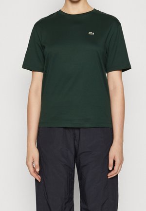 Person trägt ein dunkelgrünes T-Shirt mit kurzen Ärmeln und einem kleinen Krokodil-Logo auf der linken Brust sowie schwarze Hosen, steht vor einem schlichten Hintergrund.