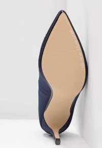 Chaussure à talon haut bleu marine à bout pointu présentée semelle vers le haut, semelle beige avec l’inscription « Life is a Showcase! » près de la pointe, chaussure devant un mur blanc.