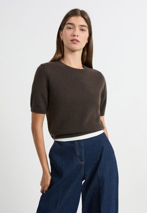 Maglione marrone a maniche corte con scollo rotondo, realizzato in materiale morbido, abbinato a jeans denim blu a gamba larga con una fascia in vita bianca.