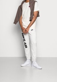 Białe dresy Nike Air z czarnym logo, zestawione z kremową koszulą i brązową bluzą narzuconą na ramiona. Całość dopełniają białe sneakersy.