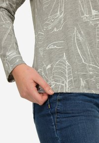 Szara, longsleeve koszula z białymi wzorami żaglówek, zestawiona z ciemnoniebieskimi dżinsami. Dłoń trzymająca brzeg koszuli, podkreślająca teksturę materiału.