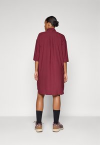 Vestido camisero color marrón oscuro con cuello, mangas cortas y un corte relajado, que presenta un pliegue en la parte trasera central y aberturas laterales para facilitar el movimiento.