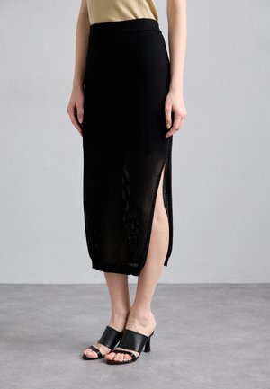 Jupe en maille noire avec fentes latérales et taille ajustée ; associée à des mules à talons blocs noirs. Tissu lisse et texturé, design longueur cheville.