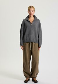 Donna in piedi che indossa un maglione grigio con zip, pantaloni marroni larghi con coulisse e ballerine in rete nera, davanti a uno sfondo chiaro e semplice.
