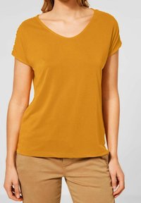 Kvinna med senapsgul kortärmad V-ringad tröja och ljusbruna byxor, stående med händerna avslappnade vid sidorna mot en enfärgad bakgrund.