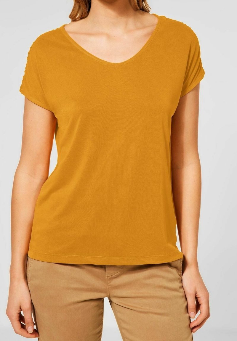 Kvinna med senapsgul kortärmad V-ringad tröja och ljusbruna byxor, stående med händerna avslappnade vid sidorna mot en enfärgad bakgrund.