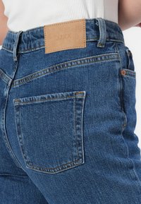 Närbild av en person som bär blå denimjeans med en brun läderloggopatch och synlig söm på bakfickan.