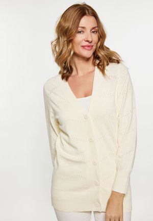 Frau mit schulterlangem, welligem blondem Haar, die eine cremefarbene, geknöpfte Strickjacke über einem weißen Oberteil vor einem einfarbigen hellen Hintergrund trägt.