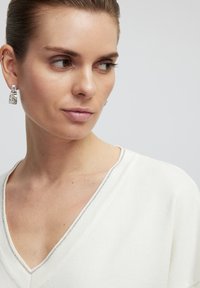 Bílý svetr s výstřihem do V a texturovaným povrchem, s jemným stříbrným lemem podél výstřihu. Modelka má na sobě výrazné stříbrné náušnice.