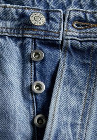 Tejido de denim azul claro con una bragueta, que presenta tres botones a presión metálicos y costuras naranjas en contraste a lo largo de las costuras.
