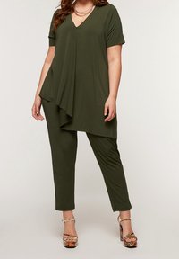 Nainen, joka on pukeutunut oliivinvihreään epäsymmetriseen tunikaan, joka on yhdistetty samanvärisiin housuihin ja korkokenkiin, joissa on kuviota, seisoo yksinkertaista taustaa vasten.