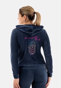 Sudadera con capucha azul marino de textura acanalada, con un diseño de piña bordado y el texto "Pineapple Bay" en rosa.