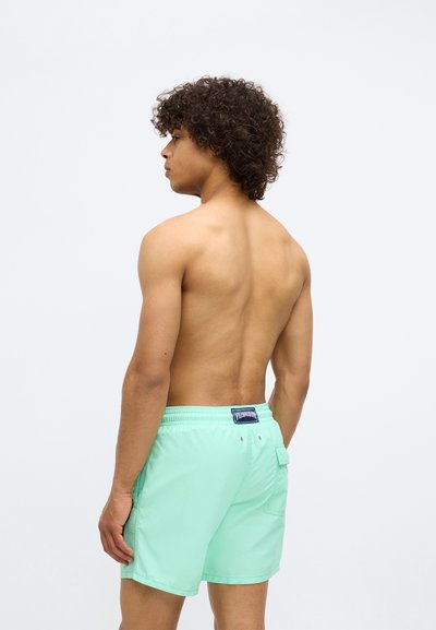 Νέος άνδρας με σγουρά μαλλιά, φορώντας μαντίλι κολυμβητηρίου σε μέντα απόχρωση, στέκεται χωρίς μπλούζα γυρίζοντας προς τα αριστερά σε έναν απλό φωτεινό φόντο.