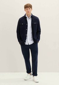 Tumesinine jakk nööpidega ja kahe esikotiga, mille juurde on kombineeritud valge nööbitav särk ja tumedad püksid, lõpetatuna valgete kingadega.