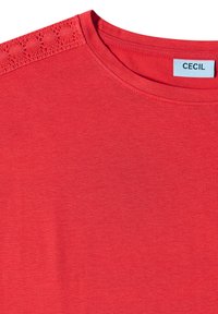 T-shirt rouge à encolure ronde avec un détail en crochet à l'épaule, montrant une étiquette blanche avec le nom de la marque "Cecil."