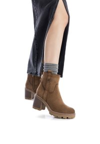 Botines de ante marrones con un tacón grueso, que presentan una punta redondeada, costuras en contraste y una suela de goma texturizada. Combinados con calcetines grises.