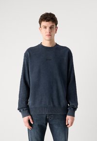 Granatowy sweter z teksturowanymi rękawami, ściągaczami i okrągłym dekoltem. Posiada dyskretną grafikę na piersi oraz luźny krój.