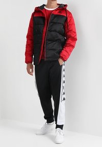 Zwarte en rode pufferjack met capuchon, ritsluiting; gecombineerd met zwarte trainingsbroek met een witte zijkant en logo. Witte sneakers.