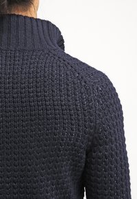 Osoba oblečená v tmavě modrém silném pleteném svetru s vysokým žebrovaným límcem, zobrazená zezadu a oříznutá v oblasti krku.