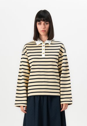 Kvinne med rett, mørkt hår, iført en beige og svartstripet skjorte med lange ermer, hvit krage og gullknapper, kombinert med et mørkt skjørt.