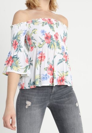 Haut blanc à épaules dénudées avec un motif floral en rouge, rose, jaune et vert. Présente un corsage smocké et des manches évasées. Associé à un jean gris déchiré.