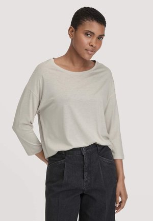 Helles beige Langarmoberteil mit weitem Ausschnitt und überschnittenen Schultern, kombiniert mit hochgeschnittenen dunkelblauen Jeans mit vorderen Falten.