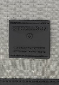 Svart gummimärke med upphöjd text "STRELLSON" och hashtag, på en texturerad grå tygbakgrund. Inkluderar geografiska koordinater.