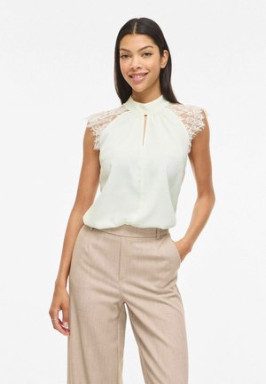 Femme portant un chemisier blanc sans manches avec des épaules en dentelle et un pantalon beige taille haute, debout avec une main dans la poche, souriante.