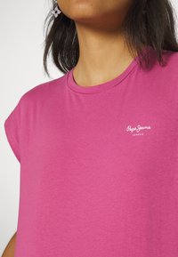 Fuksijos spalvos marškinėliai su apvaliu kaklu ir trumpomis, suktomis rankovėmis. Priekyje, kairėje pusėje, yra balta „Pepe Jeans London“ logo.