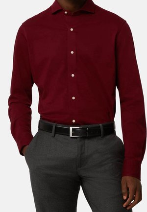 Mężczyzna w bordowej koszuli na długi rękaw, wpuszczonej w szare spodnie z czarnym skórzanym paskiem, ręce w kieszeniach.
