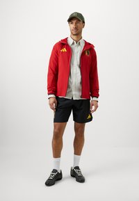 Homem vestido com um boné verde, jaqueta vermelha com fecho, camisa branca, calções pretos com logos, meias brancas e ténis pretos, de pé contra um fundo branco.