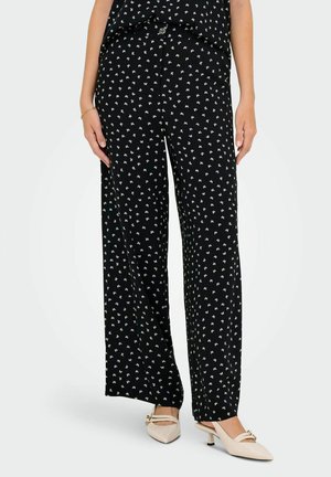 Pantalons larges noirs avec un motif floral blanc, dotés d'un tissu lisse et d'une coupe décontractée. Associés à des talons de couleur claire.