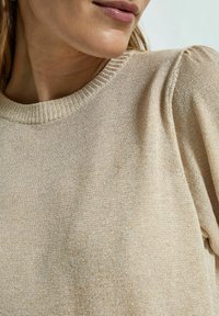 Beżowa dzianinowa bluza z prążkowanym, okrągłym dekoltem i subtelnym połyskiem. Posiada bufiaste rękawy i miękką, teksturowaną tkaninę.