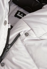 Weiße Steppjacke mit glatter Textur, ausgestattet mit metallischen Druckknöpfen und Reißverschluss. Innenetikett trägt die Aufschrift "BAKER by TEAM."