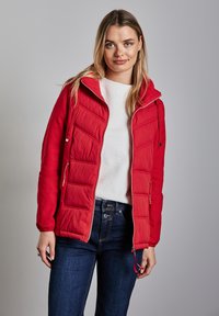 Röd vadderad jacka med dragkedja fram, justerbar huva och två sidofickor. Bärs över en vit texturerad topp och mörka denimjeans.