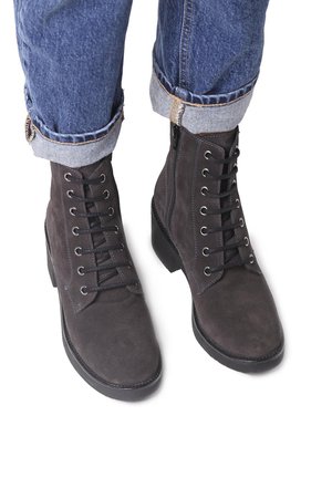 Botas de tobillo gris oscuro hechas de ante, con cordones en la parte delantera, cremallera en el costado y suela plana. Se ven pantalones de mezclilla con puños.