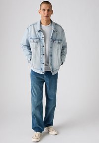 Jachetă din denim de culoare albastru deschis, cu două buzunare pe piept, un hanorac gri dedesubt, blugi largi albaștri și adidași bej. Aspect simplu și casual.