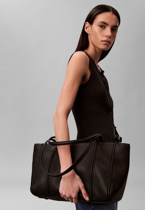 Γυναίκα με μακριά σκούρα μαλλιά που φοράει αμάνικο μαύρο τοπ κρατάει μια μεγάλη μαύρη δερμάτινη τσάντα ώμου μπροστά σε γκρι φόντο.