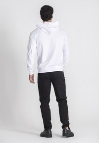 Sudadera blanca con capucha ajustable, dobladillo y puños acanalados, combinada con jeans negros y zapatos negros, vista desde la espalda.