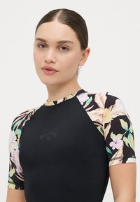 Traje de neopreno de manga corta en negro con mangas de estampado floral. Presenta un cuello alto y un detalle de logo en el centro del pecho.